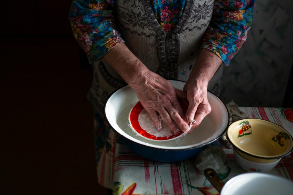 A senior woman washing a red dish in a vintage kitchen setting with floral patterns.